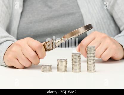 Eine Frau sitzt an einem Tisch und hält eine hölzerne Lupe über Stapel weißer Münzen. Das Konzept der Budgetplanung, Einsparungen. Investitionssuche Stockfoto