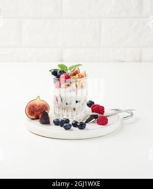 Ein transparentes Glas mit Müsli, gegossen mit Joghurt, auf einem weißen Tisch mit reifen Himbeeren, Blaubeeren und Feigen. Gesundes Frühstück Stockfoto
