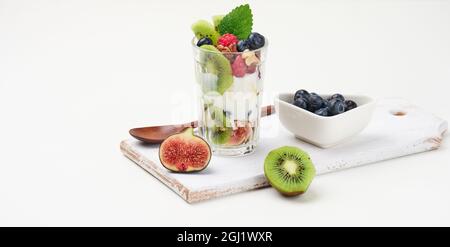 Ein transparentes Glas mit Müsli, gegossen mit Joghurt, auf einem weißen Tisch mit reifen Himbeeren, Blaubeeren und Feigen. Gesundes Frühstück Stockfoto