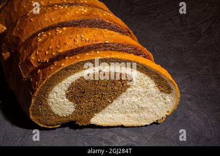 Ungewöhnlicher, frischer zweifarbiger Laib Brot auf schwarzem Hintergrund, Foto in einem niedrigen Ton bei hartem Licht, Nahaufnahme. Stockfoto