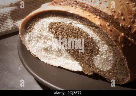 Ungewöhnliches, zweifarbiges frisches Brot in einer Keramikplatte und eine gewebte Serviette auf schwarzem Hintergrund, Foto in einem niedrigen Ton bei hartem Licht, Nahaufnahme. Stockfoto