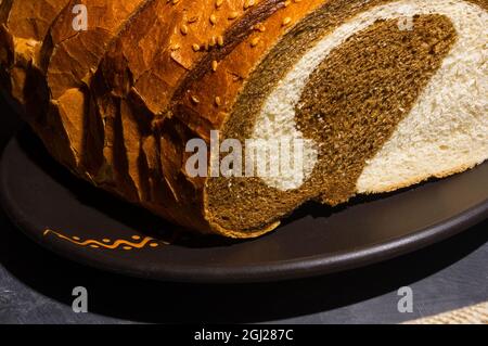 Ungewöhnliches, zweifarbiges frisches Brot in einer Keramikplatte und eine gewebte Serviette auf schwarzem Hintergrund, Foto in einem niedrigen Ton bei hartem Licht, Nahaufnahme. Stockfoto