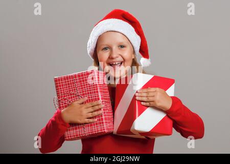 Glückliches kleines Mädchen hält zwei Boxen mit Weihnachtsgeschenken isoliert auf grauem Hintergrund. Ein Kind in einem Weihnachtsmann lacht und schaut auf die Kamera. Stockfoto