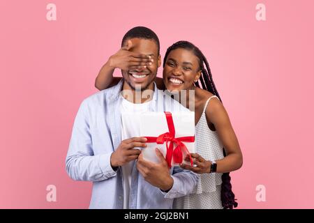 Konzept Der Feier. Eine aufgeregte afroamerikanische Dame, die die Augen ihres Freundes bedeckt, die verpackte Geschenkbox hält und ihn mit Geburtstag oder Jubiläum begrüßt Stockfoto