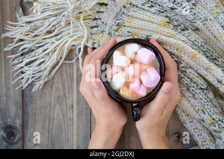 Weibliche Hände halten Becher mit heißer Schokolade und Marschwalben in Herzform. Wärmendes Getränk auf Holztisch und Strickschal. Weicher Fokus Stockfoto