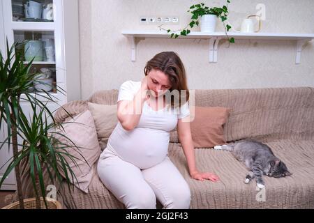 Eine Schwangere hält sich an einem schmerzenden Hals fest, während sie auf einem Sofa im Zimmer sitzt Stockfoto