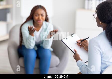Depressive schwarze Frau, die Psychologin konsultiert, negative Emotionen in der psychiatrischen Klinik zum Ausdruck bringt, Raum kopieren. Psychotherapeutin hilft unglücklichen Afro Dame wi Stockfoto
