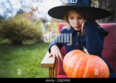 Gruseliges kleines Mädchen im Hexenkostüm, Hut mit großem Kürbis, der Halloween-Urlaub feiert. Sitzend auf Sessel im Mantel mit Kürbis. Stilvolles Bild. Horro Stockfoto