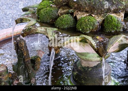 Nahaufnahme des fließenden Wassers aus dem oberen Stockwerk des Steingutbrunnens mit den vielen Flechten zum unteren Stockwerk, dekoriert im Garten des Stockfoto