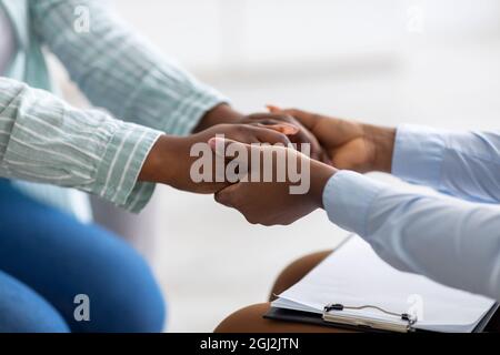 Nahaufnahme einer Psychotherapeutin, die depressive junge schwarze Frau unterstützt, ihre Hände in der psychiatrischen Klinik haltend, abgeschnitten. Nicht erkennbarer Psychologe hilft st Stockfoto