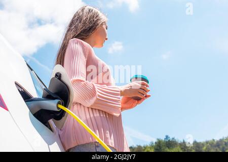 Eine Frau, die eine Kaffeepause nimmt, während sie ihr Elektroauto lädt Stockfoto
