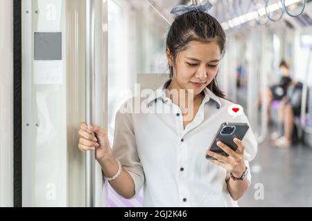 Junge asiatische Frau, die ein Mobiltelefon mit Herz-Symbol in einer leeren U-Bahn oder einem Skytrain benutzt Stockfoto