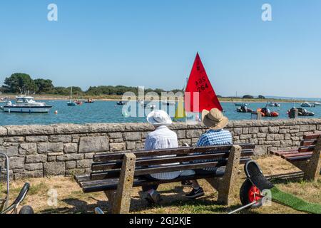 Seniorenpaar sitzt auf einer Bank und beobachtet Boote und Schlauchboote beim Segeln in Keyhaven, Hampshire, England, Großbritannien Stockfoto