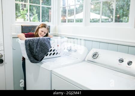 Ein kleines Mädchen mit langen Haaren und Sommersprossen hält einen Wäschekorb und macht die Wäsche als Waschküche in einem natürlichen hellen Waschraum mit Waschmaschine Stockfoto