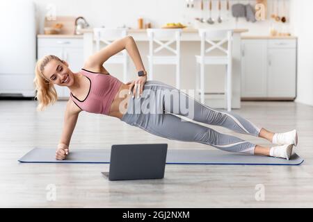 Lächelnde europäische junge blonde Fitness-Dame macht Seitenwand und sehen Online-Trainingsstunde auf Laptop zu Hause. Abnehmen, Sport aus der Ferne, fit bleiben Stockfoto
