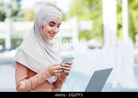 Beschäftigt lächelnd niedlich tausendjährige arabische muslimische Dame in Hijab tippen auf dem Smartphone, verwenden Laptop im Freien. Geschäftsfrau arbeitet oder studiert in der Stadt mit Geräten, f Stockfoto