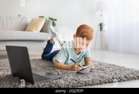 Smart Kid Ingwer Junge Schuler auf Teppich mit Laptop legen, Notizen machen, Hausaufgaben machen, Surfen im Internet oder Online-Klasse, Wohnzimmer i Stockfoto