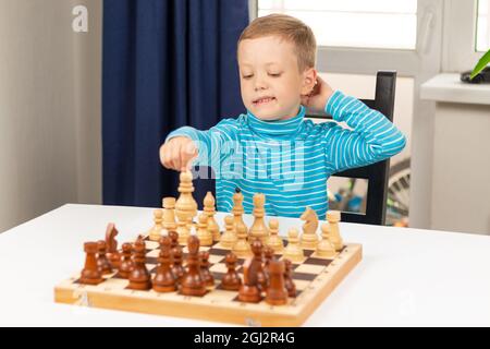 Netter siebenjähriger Junge, der zu Hause auf einem weißen Holztisch Schach spielt. Selektiver Fokus. Nahaufnahme. Hochformat Stockfoto