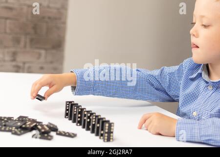 Der sechsjährige niedliche Junge spielt zu Hause auf einem weißen Holztisch Dominos. Selektiver Fokus. Nahaufnahme Stockfoto