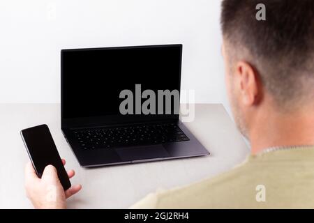 Ein Mann mit einem Telefon in der Hand sitzt vor einem Laptop mit einem schwarzen Mock-up auf dem Monitor, das Konzept der Büroarbeit, Remote-Arbeit, Marketing, Trai Stockfoto