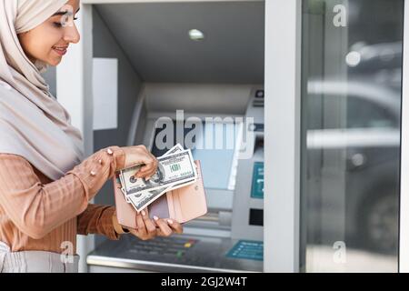 Lächelnde junge arabisch-islamische Geschäftsfrau in Hijab legt Dollar in Brieftasche neben geldautomaten mit leerem Bildschirm. Lastschrift, Online-Guthaben, Barauszahlung, m Stockfoto