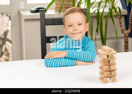 Ein süßer siebenjähriger Junge, der zu Hause an einem weißen Holztisch vor dem Hintergrund eines Lichtfensters Jenga spielt. Selektiver Fokus. Nahaufnahme. Por Stockfoto