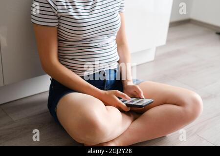 In der modernen Küche sitzt die junge Frau mit einem Smartphone auf dem Boden. Frau schreibt mit ihrem Freund. Freiberuflicher Mitarbeiter, der zu Hause arbeitet Stockfoto