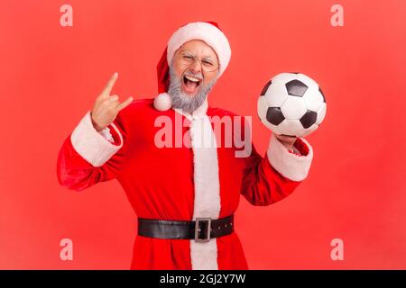 Aufgeregt verrückter älterer Mann mit grauem Bart trägt weihnachtsmann Kostüm steht mit Fußball in den Händen und zeigt Felsschild vor der Kamera. innenstudi Stockfoto