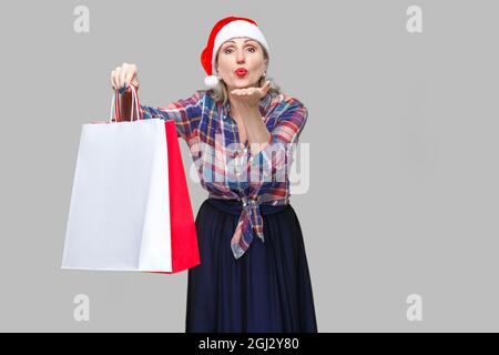 Portrait von bitte schön erwachsenen Frau in rot santa Kappe und kariertem Hemd, die hielten Einkaufstaschen und senden Sie küssen, am Cam suchen Stockfoto