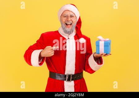 Extrem glücklicher älterer Mann mit grauem Bart trägt weihnachtsmann Kostüm hält Geschenk-Box und zeigt auf Geschenk mit positiver Optik. Studio-Sho Stockfoto