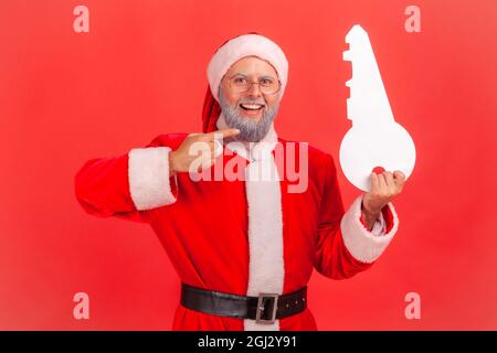 Portrait eines lächelnden älteren Mannes mit grauem Bart, der das weihnachtsmann-Kostüm trägt, der auf den Papierschlüssel zeigt, empfehle die Agentur für den Kauf einer Wohnung. Ind Stockfoto