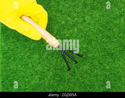 Ansicht einer menschlichen Hand in einem gelben Handschuh, der einen Handgrubber vor einem grünen Rasen hält Stockfoto