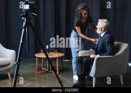 Brunette afroamerikanische Assistentin Anpassung Mikrofon auf Anzug von Geschäftsmann im Interview Studio Stockfoto