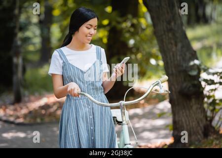 Erfreut asiatische Frau mit Fahrrad Messaging auf Smartphone im Park Stockfoto