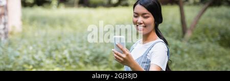 Fröhliche asiatische Frau, die im Park auf dem Handy chattet, Banner Stockfoto