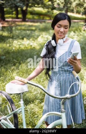 Positive asiatische Frau mit Kopfhörern chattet auf dem Handy in der Nähe des Fahrrads im Park Stockfoto