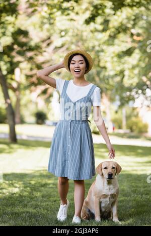 Aufgeregt asiatische Frau in gestreiften Sundress und Strohhut Blick auf Kamera in der Nähe Hund im Park Stockfoto