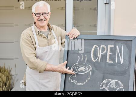 Porträt eines glücklichen, reifen Besitzers der Bäckerei mit Schnurrbart, der beim Eingang steht und auf eine Tafel mit offenem Schild zeigt Stockfoto