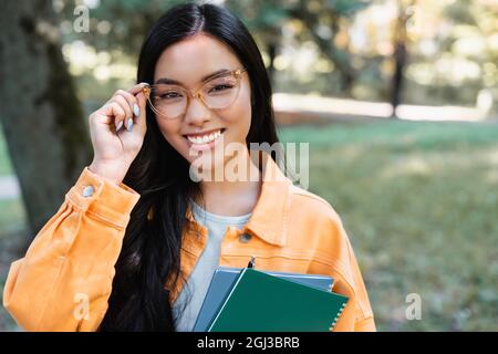 Brunette asiatische Student Anpassung Brillen, während die Notizbücher im Park Stockfoto