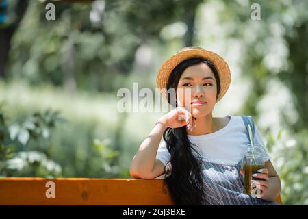 Verträumte und lächelnde asiatische Frau im Strohhut sitzt mit frischem Smoothie im Park Stockfoto