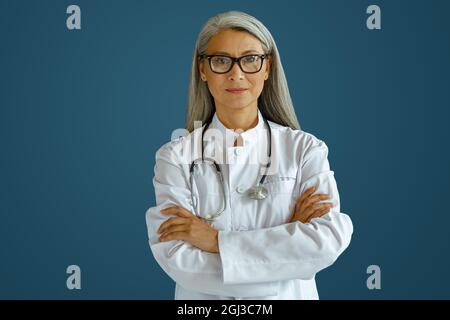 Reife Ärztin in Robe mit Brille und Stethoskop steht auf blauem Hintergrund Stockfoto