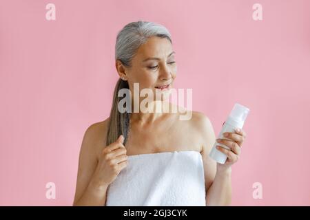 Schöne reife asiatische Dame mit Handtuch verpackt hält Flasche Körperpflegeprodukt im Studio Stockfoto