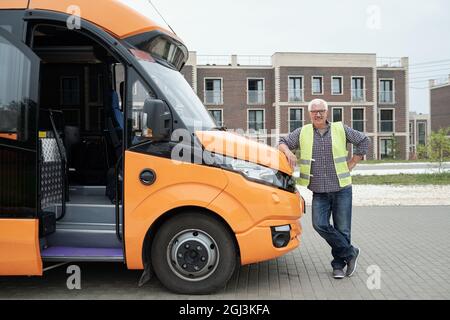 Lächelnder, alter kaukasischer Busfahrer in Brillen und grüner Weste, der sich auf die Motorhaube stützt, während er auf Passagiere wartet Stockfoto