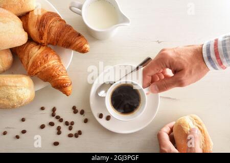 Mann beim Frühstück mit einer Tasse Kaffee, Teller mit Gebäck, Butter und Milch auf einem weißen Holztisch. Draufsicht. Horizontale Zusammensetzung. Stockfoto