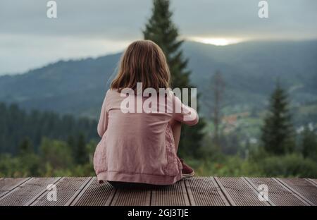 Kleines blondes Mädchen, das auf der Terrasse sitzt und den Sonnenuntergang hinter den Bergen betrachtet Stockfoto