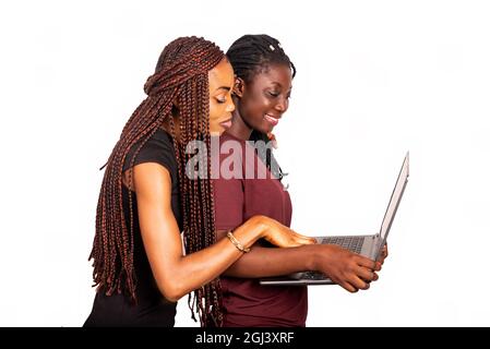 Zwei wunderschöne weibliche Studenten stehen auf weißem Hintergrund und arbeiten mit einem Laptop, während sie lächeln. Stockfoto