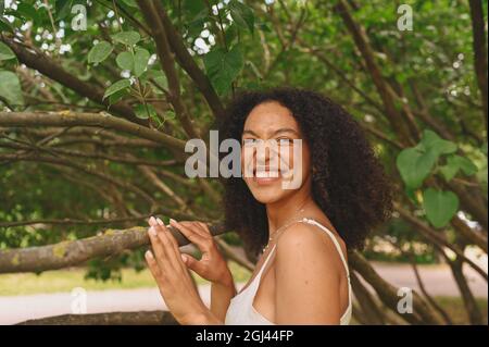 Mode Nahaufnahme Porträt des Lachens Attraktive junge natürlich schöne afroamerikanische Frau mit afrohaaren und perfekten Zähnen Lächeln posiert in Natur Parkland in grünem Laub Stockfoto