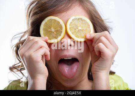 Verschmitztes kleines Mädchen, das geschnittene Zitronen an die Augen hält, während sie ihre Zunge an der weißen, isolierten Kamera ausstreckt Stockfoto