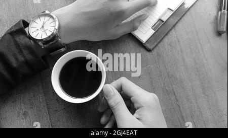 WONOSOBO, INDONESIEN - 07. Apr 2021: Eine Draufsicht von einer Person, die eine Tasse schwarzen Kaffee auf einem Holztisch in Graustufen genießt Stockfoto