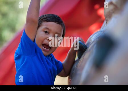 Ein netter Junge, der vor Freude schreit, als er auf die Spitze einer Outdoor-Kletterwand für Kinder klettert. Stockfoto